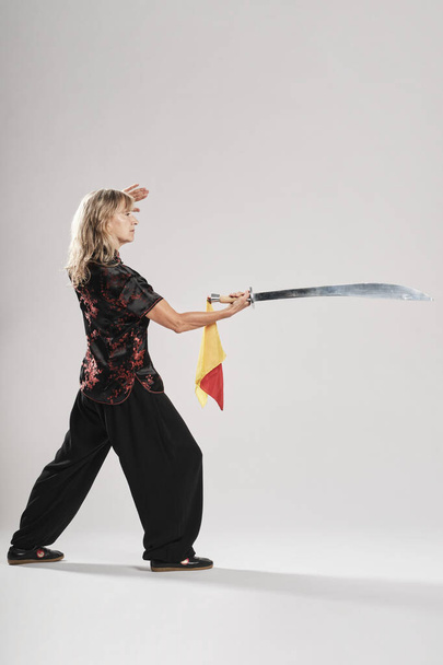Reife blonde Frau, die Tai Chin mit Säbel vor weißem Hintergrund praktiziert, trägt eine traditionelle chinesische schwarze Jacke mit roter chinesischer Dekoration, schwarze Hosen und chinesische Schuhe mit Ying Yang Symbol - Foto, Bild