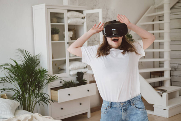 Young woman in jeans and white T-shirt wearing virtual reality helmet plays game, works, watches video in room next to bed. Concept of modern technology, VR, augmented virtual games, entertainment. - Photo, image