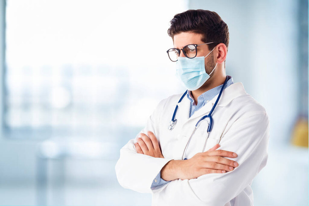 Portrait of male doctor wearing face mask while standing with arms crossed at hospitals foyer - Фото, изображение