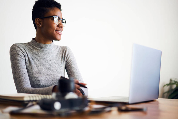 Positive black female freelancer wearing casual clothes and eyeglasses looking away while working at table with laptop and photo camera - Fotó, kép