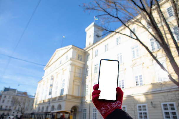 mano de mujer en guantes rojos sosteniendo el teléfono con pantalla blanca Ayuntamiento de Lviv en el concepto de viaje espacial copia de fondo - Foto, imagen