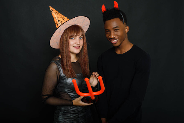 黒人青年と白人女性は黒を背景に衣装を着たスタジオでハロウィンを祝う。笑顔の若い友達 - 写真・画像