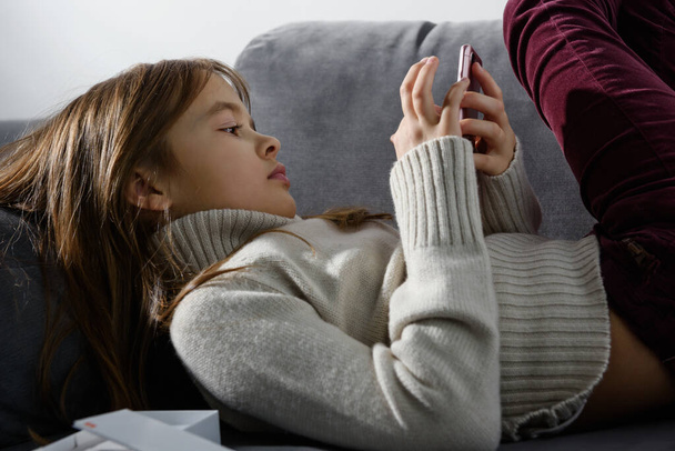 Das junge Mädchen blickt auf den Bildschirm eines Smartphones, das auf einem Sofakissen lehnt. - Foto, Bild