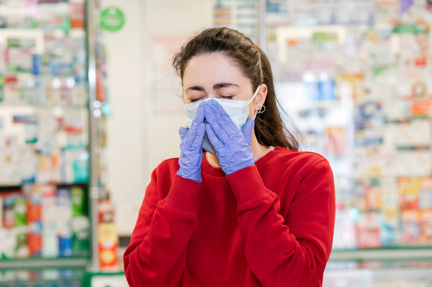 A young woman in a medical mask and gloves sneezes, covering her face. In the background, the shop Windows of pharmacy products. Concept of disease, virus protection and pharmacy business. - Photo, Image