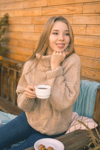 Autumn mood. A smiling girl in a cozy brown sweater and blue jeans sits on a bench and drinks coffee with croissants - Foto, afbeelding