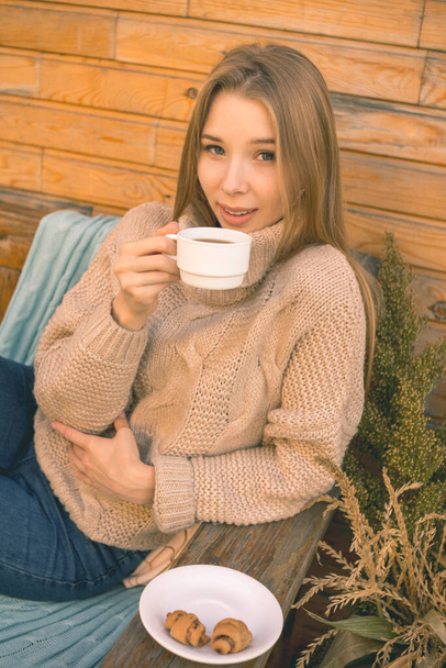 Autumn mood. A smiling girl in a cozy brown sweater and blue jeans sits on a bench and drinks coffee with croissants - Fotografie, Obrázek
