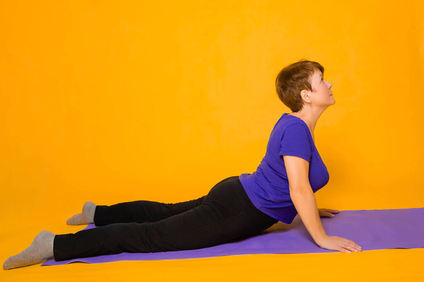 Aged woman doing yoga on a purple rug. Studio photo on a yellow background. - Fotoğraf, Görsel
