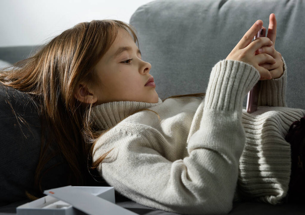 Το νεαρό κορίτσι κοιτάζει την οθόνη ενός smartphone ακουμπισμένο σε ένα μαξιλάρι καναπέ. - Φωτογραφία, εικόνα