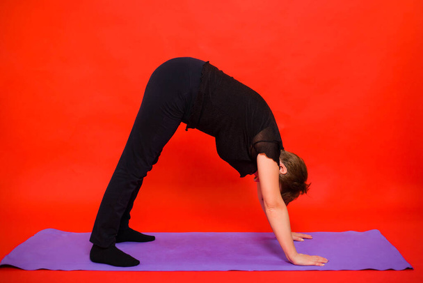 Aged woman in black clothes doing yoga on a purple rug. Studio photo on a red background.  - Fotografie, Obrázek