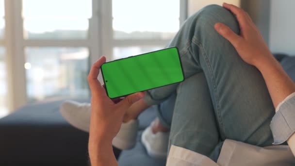 自宅の男性はソファに横になり、水平モードで緑色のモックアップ画面を持つスマートフォンを使用しています。彼はインターネットを閲覧し、コンテンツ、ビデオ、ブログを見て - 映像、動画