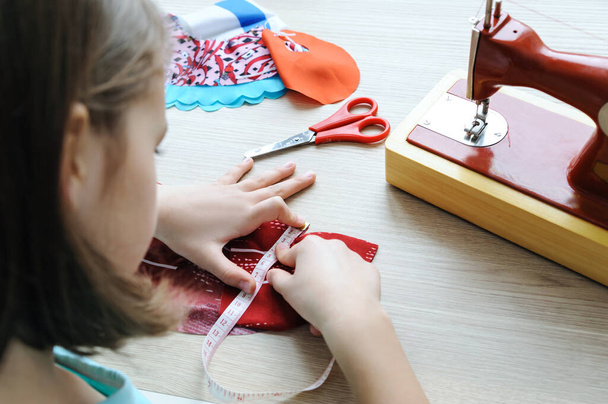 Tyttö ompelee pukua nukelle. Hän käyttää mittanauhaa tuotteen mittaamiseen.. - Valokuva, kuva