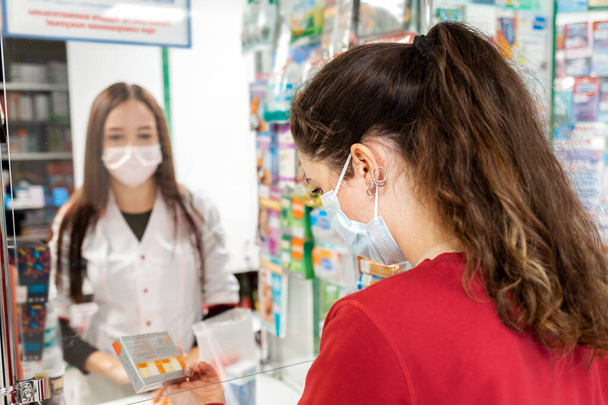 A brunette woman in a medical mask reads the composition of the drug, rear view. In the background, a pharmacist in a blur. The concept of buying medicines in a pharmacy. - Photo, Image