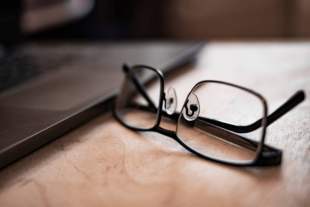 Background image of business office with laptop on wooden desk, focus on black hard rim glasses in foreground, copy space. Out of focus. Blurred - Zdjęcie, obraz