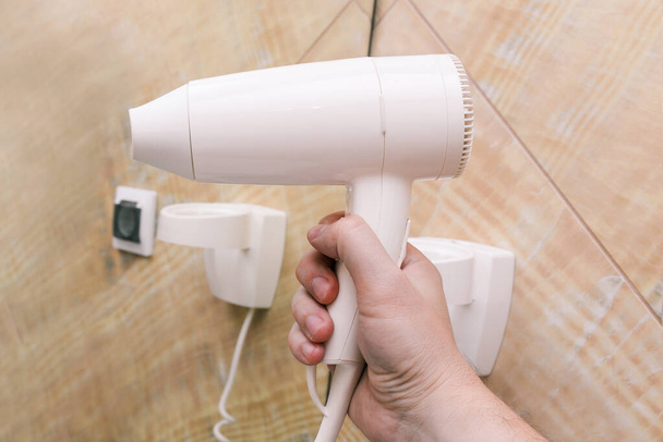 White hair dryer close-up. A man holds a plastic dryer in his hand against a background of mirrors and marble tiles. Interior of a bathroom, beauty salon or hair salon - Fotografie, Obrázek