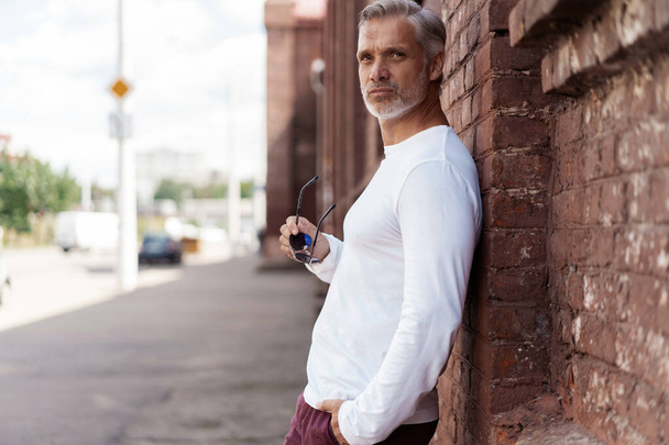 Portrait of a Grey-haired Mature handsome man in jeans and white t-shirt leaning to the old wall - Photo, image