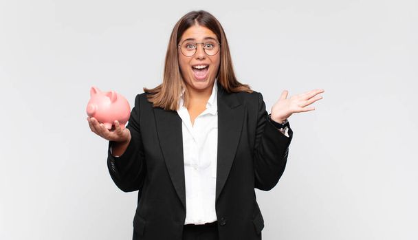 young woman with a piggy bank feeling happy, excited, surprised or shocked, smiling and astonished at something unbelievable - Foto, Imagen