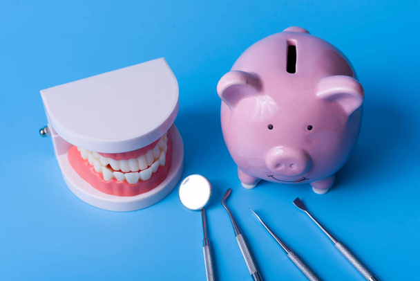 Piggy Bank na niebieskim tle i instrumenty dentystyczne. Koncepcja oszczędzania pieniędzy na produkcję implantów i leczenie stomatologiczne. - Zdjęcie, obraz
