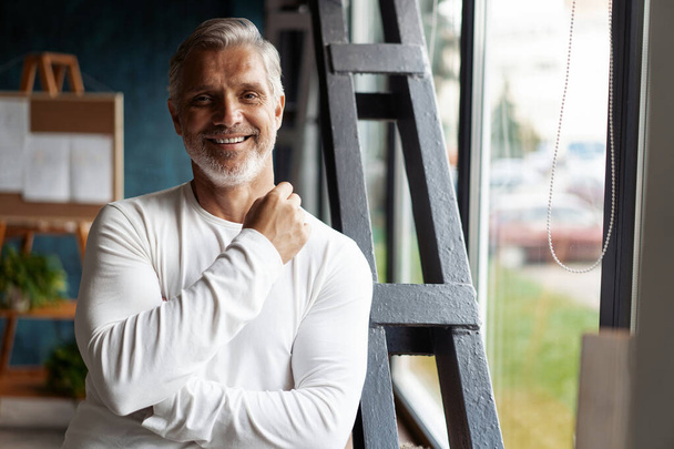 Smiling attractive Casual Grey-haired mature man standing near window looking at the camera. - Φωτογραφία, εικόνα