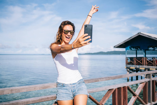 Carefree female blogger in sunglasses shooting streams vlog in live mode using roaming internet connection for web communication, happy girl making selfie photos during getaway trip to Raja Ampat - Foto, afbeelding