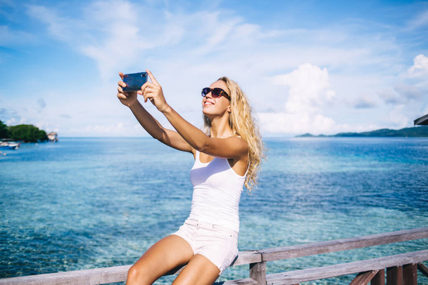 Sinceramente mujer en gafas de sol sonriendo mientras se fotografía a sí misma con el paisaje marino de Bahamas, feliz turista caucásico utilizando la cámara frontal del teléfono inteligente para hacer clic en los medios de comunicación selfie durante las vacaciones de viaje - Foto, imagen