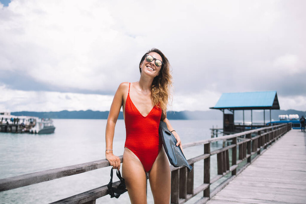 Half length portrait of cheerful Caucasian swimmer in trendy sunglasses holding snorkel mask and flippers and laughing at camera, happy female satisfied with hobby time during vacations on Bahamas - Фото, изображение