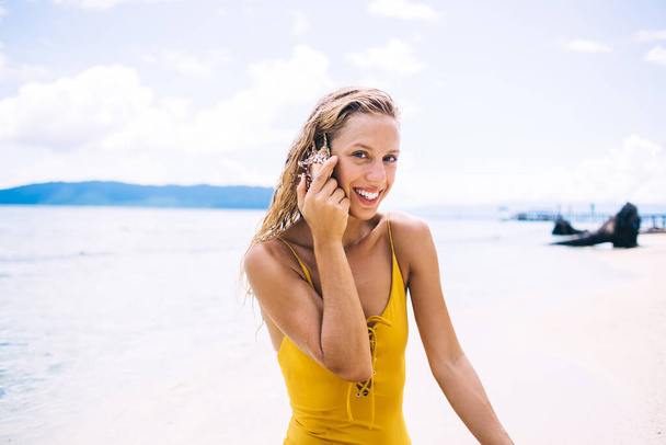 Young blonde with wet hair in yellow swimsuit sitting on beach and listening to seashell against background of sand and mountains while looking at camera - Fotoğraf, Görsel