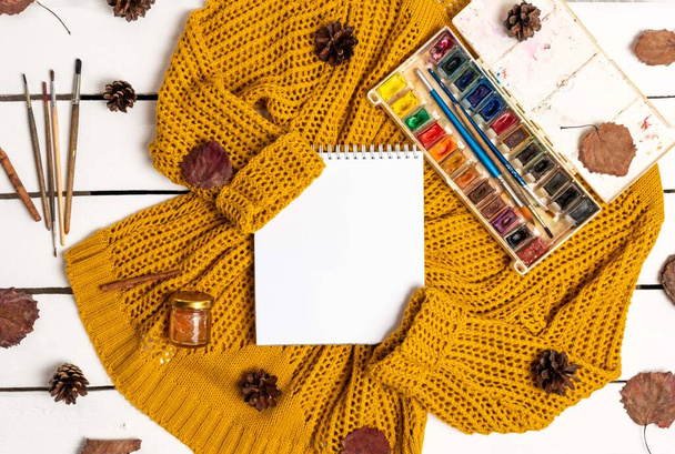 Flat lay of notebook, paints with brushes, yellow sweater, leaves, cinnamon sticks and pine cones on a white wooden background. Autumn composition with place for text - Foto, immagini