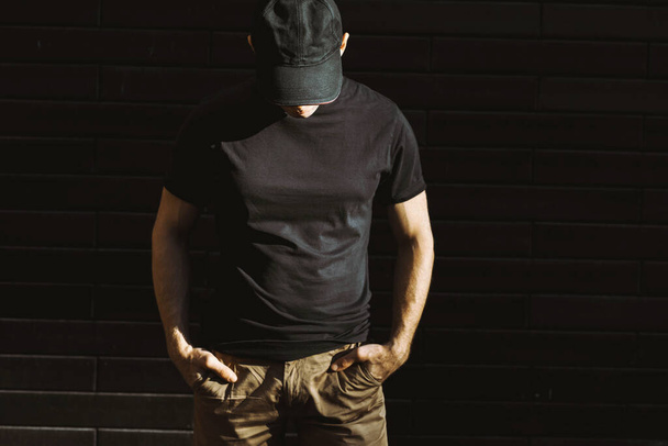 Mockup uomo in t-shirt nera e berretto da baseball in posa contro un muro nero - Foto, immagini