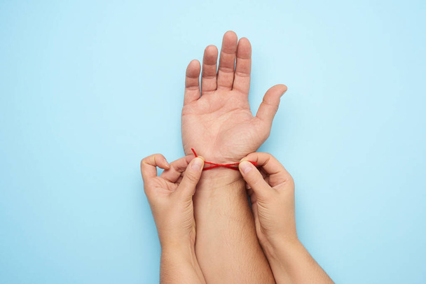 zwei weibliche Hände binden einen roten Wollfaden am Handgelenk einer männlichen Hand, blauer Hintergrund, Ansicht von oben - Foto, Bild