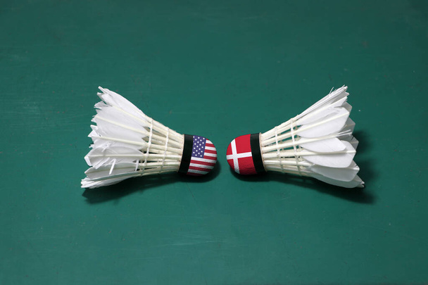 Twee gebruikte shuttlecocks op de groene vloer van Badminton Court met beide hoofden tegen elkaar. Een hoofd geschilderd met de Amerikaanse vlag en een hoofd geschilderd met de Deense vlag, concept van badminton competitie. - Foto, afbeelding
