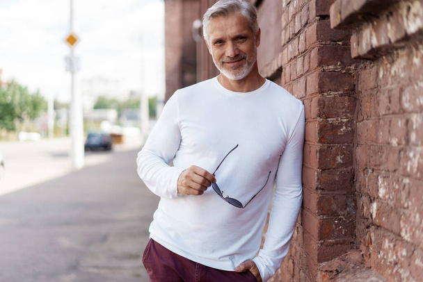 Portrait of a Grey-haired Mature handsome man in jeans and white t-shirt leaning to the old wall - Photo, image