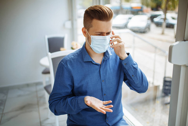 Young businessman wearing a medical sterile mask talks on the mobile phone leading business from a cafe, working remotely during coronavirus pandemic quarantine. Health safety concept - Фото, изображение