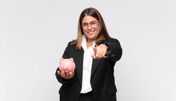 young woman with a piggy bank pointing at camera with a satisfied, confident, friendly smile, choosing you - Foto, Imagen