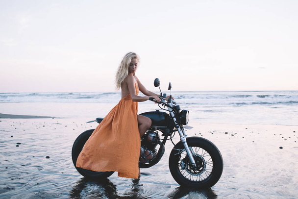 Gorgeous blond woman in long light dress posing on black vintage motorcycle on empty ocean beach in early morning looking away  - Фото, изображение