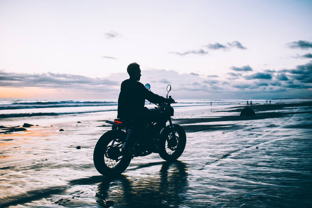 Back view of sportive man riding on motorcycle through wet beach along breathtaking dark ocean and gloomy cloudy sky - Foto, Bild