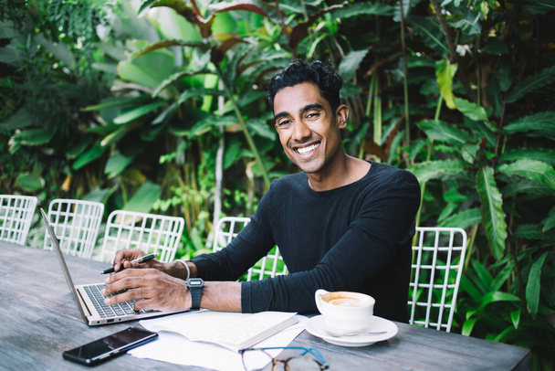 Adult Hispanic man in black shirt looking at camera and smiling while sitting at table with documents glasses and cup of hot drink and using laptop in garden during daytime on blurred background of big green tropical plants - Photo, image