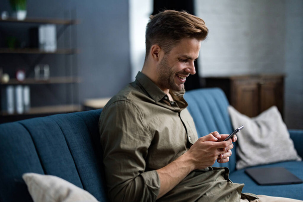 Young freelancer using smartphone and smiling. Happy man using mobile phone apps, texting message, browsing internet, looking at smartphone, sitting at home. Young people working with mobile devices - Photo, Image