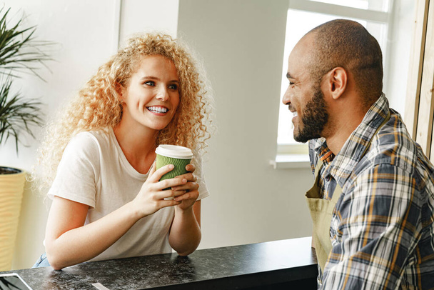 Smiling blonde woman talking to a waiter of a coffee shop at the counter - Photo, Image