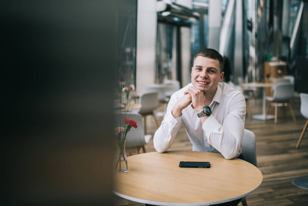 Positive male in white shirt and wristwatch sitting at wooden table with smartphone and smiling while looking at camera in cafe - Photo, image
