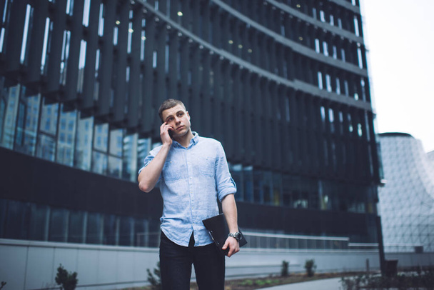 Serious young male manager in formal wear with documents in hand having phone conversation standing against contemporary building in business district - Фото, изображение