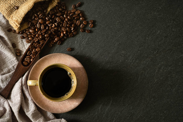 Close-up of Black hot coffee for morning with milk foam in old pink ceramic cup and napkin with coffee beans roasted in sack on dark table stone background. Top view, flat lay with copy space. - 写真・画像