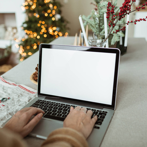 Young woman work on laptop computer with blank display screen with mockup copy space. Cozy comfortable kitchen with table, Christmas tree, fir branches, cones, candles. Holidays celebration. - Fotoğraf, Görsel
