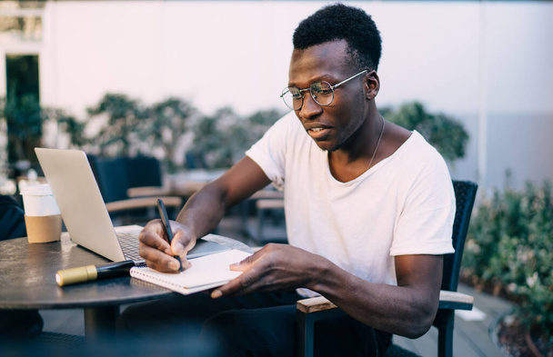 Skilled dark skinned male blogger in trendy wear planning online project making notes in notepad, creative man student learning online courses using laptop computer for searching information on terrace - 写真・画像