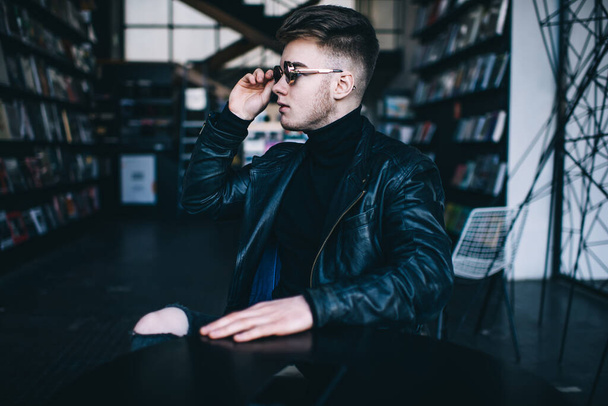 Concentrated young unshaven male with stylish haircut in black leather jacket adjusting sunglasses while sitting at table in modern cafe - Foto, immagini