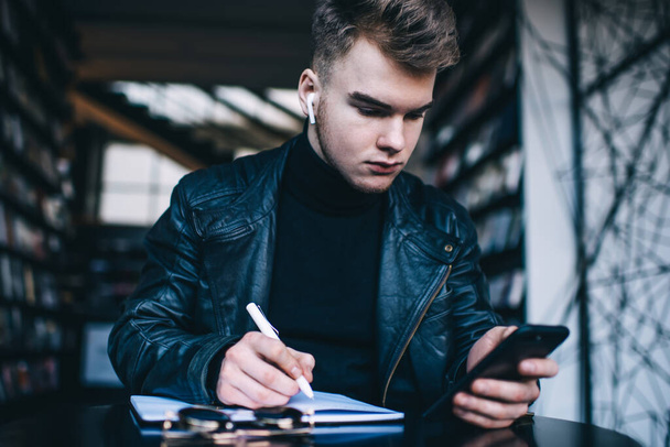 Concentrated young man in trendy casual outfit with wireless earphones using mobile phone and taking notes in notebook while sitting at table in modern library - Zdjęcie, obraz