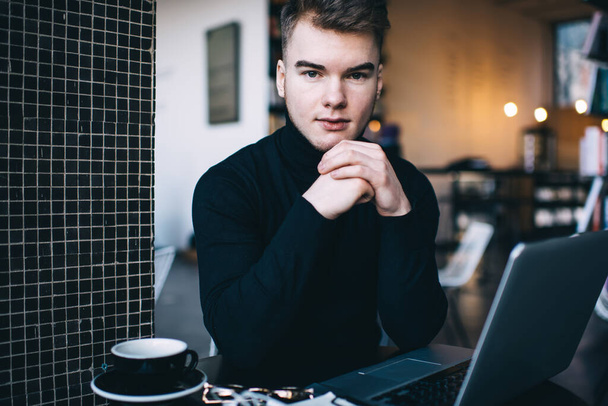 Serious young man in casual clothing sitting in coworker space in front of laptop and looking at camera while having coffee break - Zdjęcie, obraz