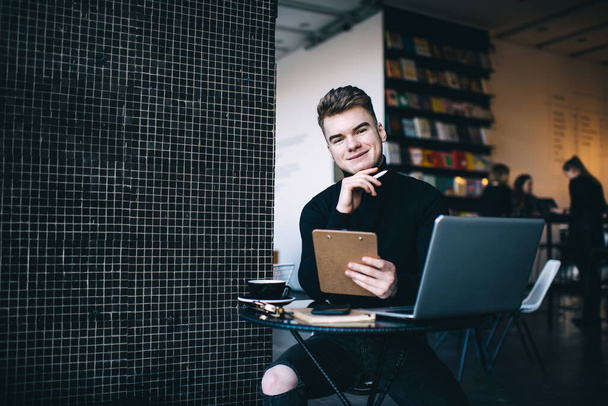 Cheerful young male student wearing black sweater looking at camera and smiling while sitting at round table with laptop and clipboard during coffee break - Foto, immagini
