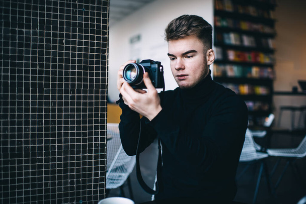 Young pensive professional male photographer in black wear taking photo on camera while standing near tile wall in library on blurred background - Valokuva, kuva