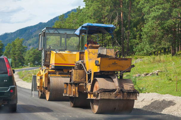 Construction machinery on the side of the road to cover asphalt - two rollers with yellow cabins at work against the backdrop of green trees and mountains as cars move. - Photo, Image