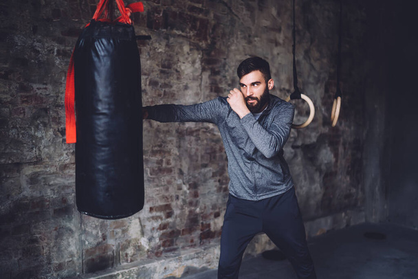 Serious handsome sportsman spending time in gym having boxing workout for training strength and coordination, young 20s caucasian man punching bag in gloves reaching body shape and slimming - Foto, Imagem